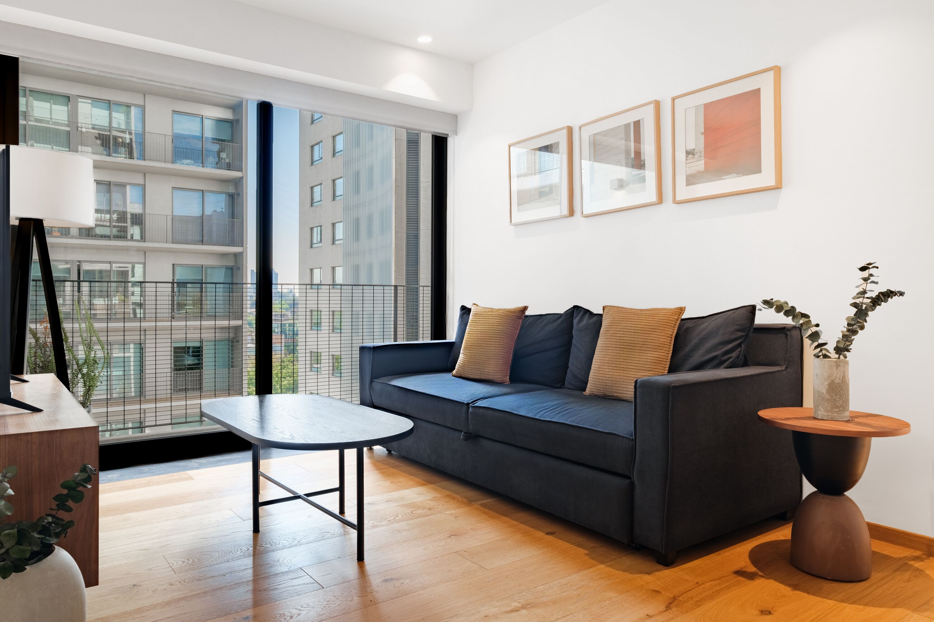 A modern living room with a dark blue sofa, a round wooden coffee table, and a small side table with a plant. There are three framed pictures on the wall and a large window offering a view of a building.