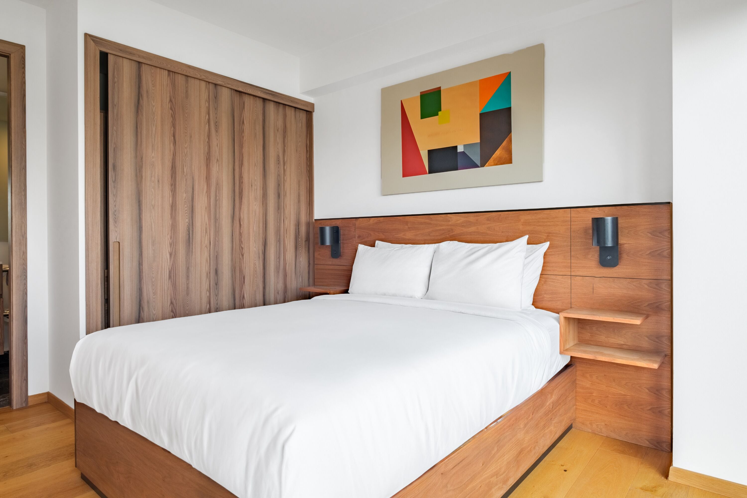 Un dormitorio moderno con estructura de cama de madera, ropa de cama blanca y una obra de arte geométrica en la pared.