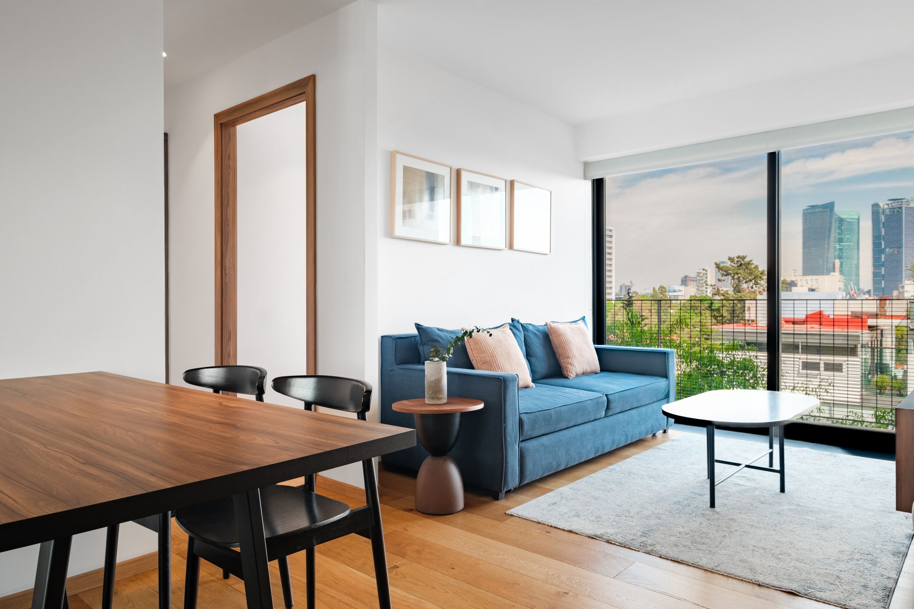 Een moderne woonkamer met een houten eettafel, een blauwe bank en een stadszicht door grote ramen.