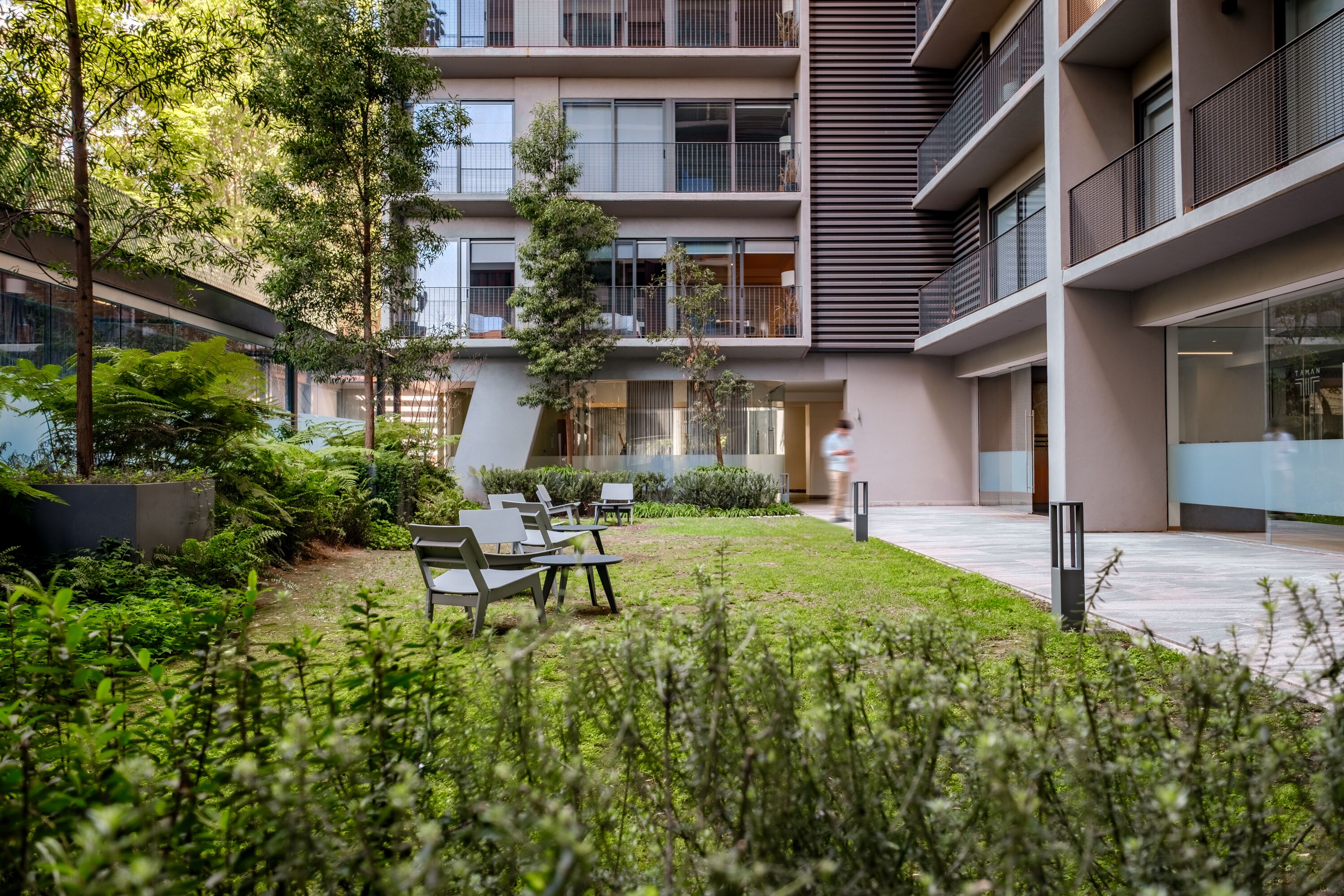 A modern outdoor courtyard with seating, greenery, and a multi-story building with balconies.