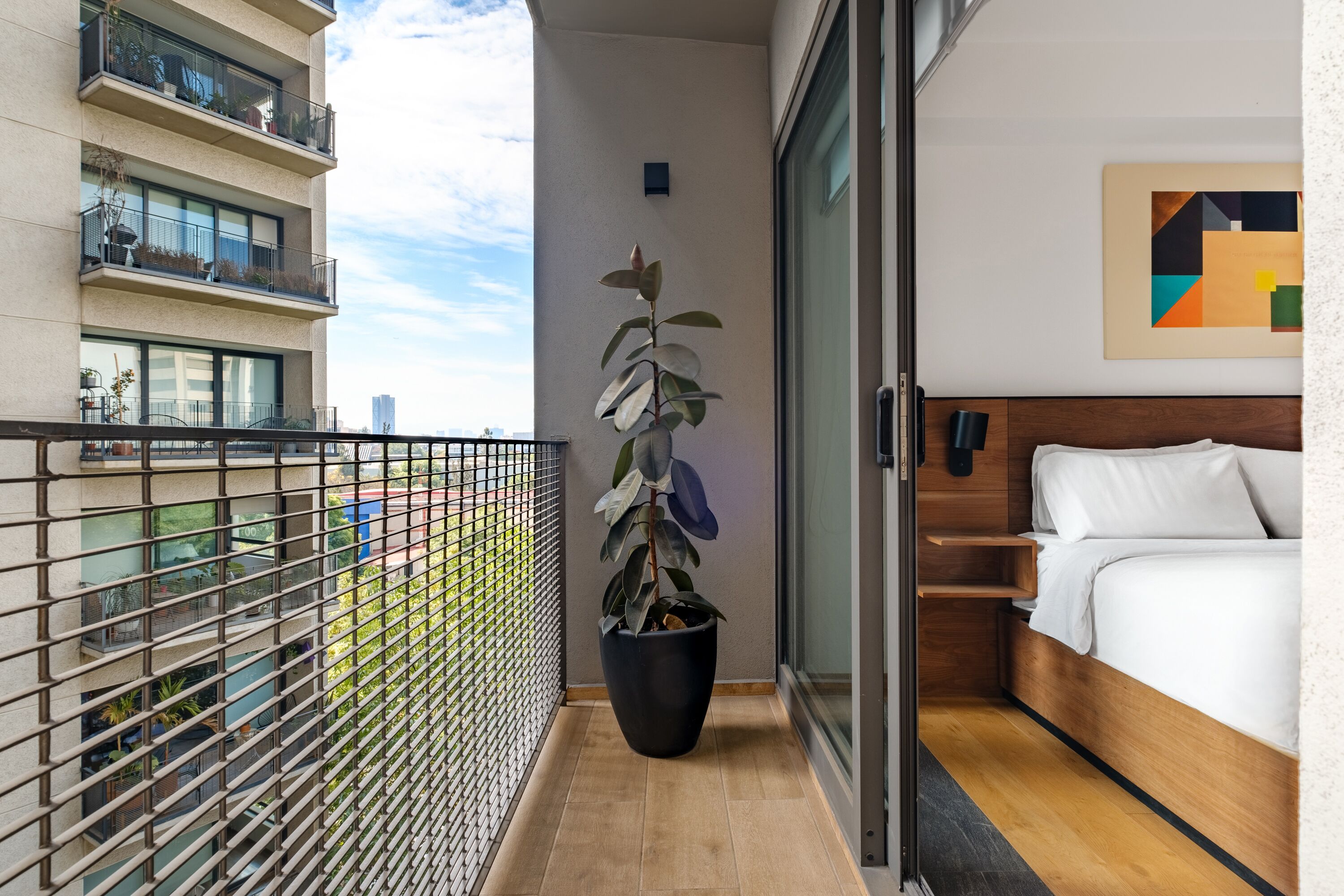 Une chambre d’hôtel avec un lit, une plante en pot, un tableau et un balcon donnant sur la ville.