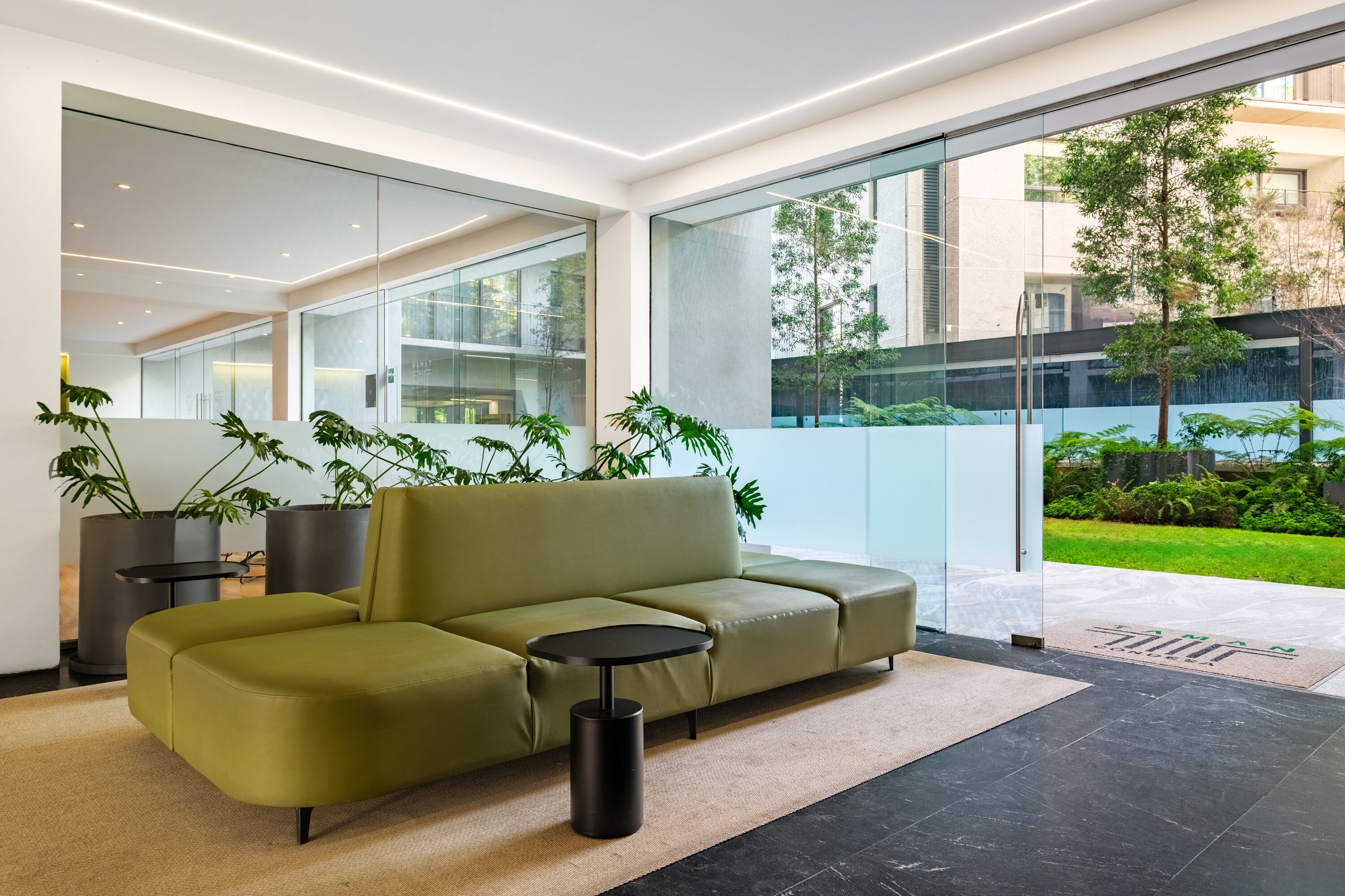 Un interior moderno con un sofá verde, una mesita redonda y grandes ventanales con vistas a un jardín.