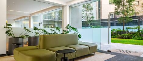 Un interior moderno con un sofá verde, una mesita redonda y grandes ventanales con vistas a un jardín.