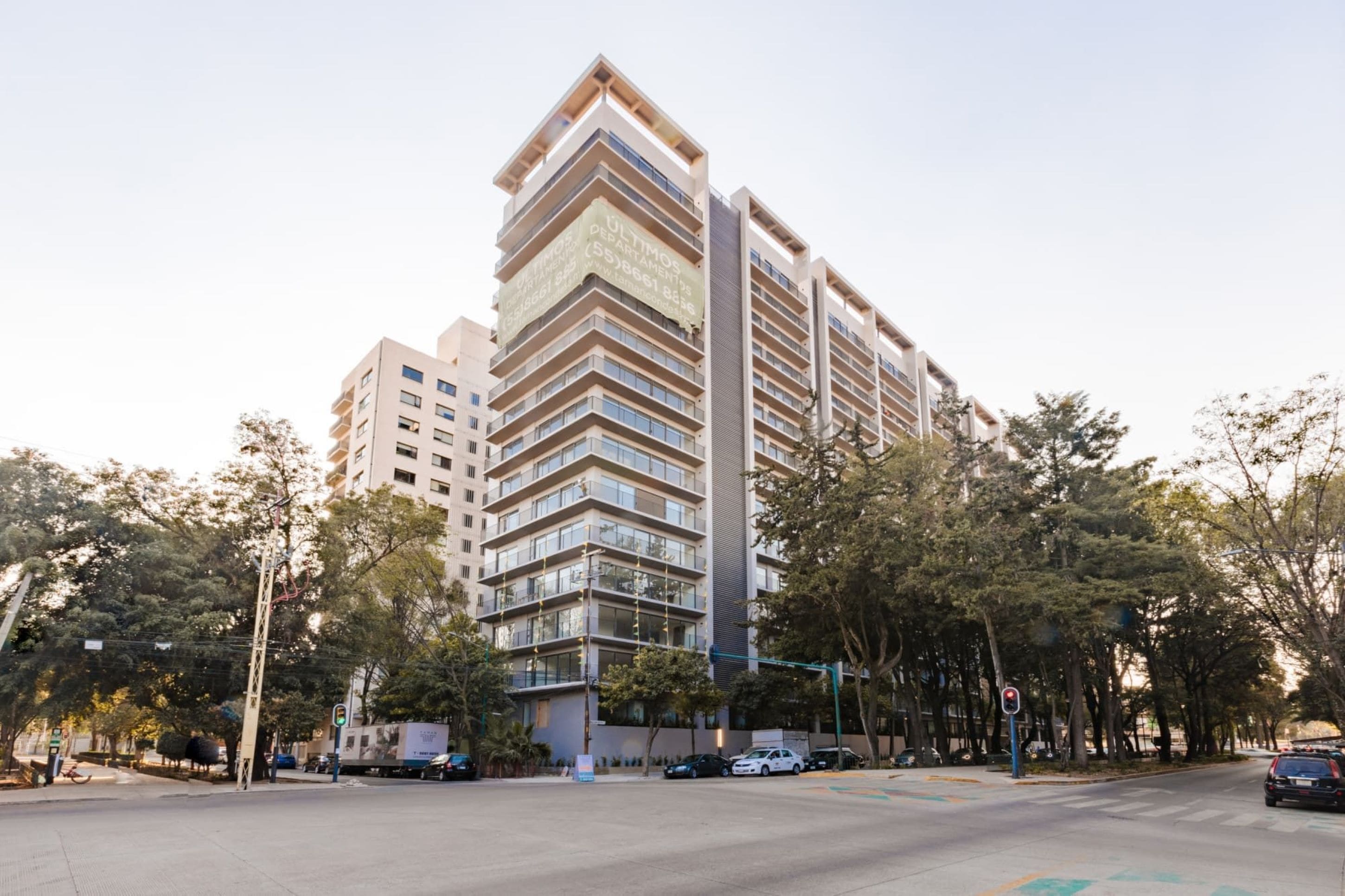 Un immeuble à plusieurs étages doté de balcons, une enseigne bien visible indiquant les coordonnées, et une rue bordée de voitures garées et d’arbres.