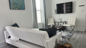 A modern living room with a white sofa, a coffee table, a TV mounted on the wall, and a window with blinds. - Serenity Pines, one bedroom, one bathroom apartment with full kitchen and garage (Pinehurst)