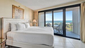 Hotelkamer met een groot bed, nachtkastje, lamp, een lijst met foto en een balkon met uitzicht op de oceaan.