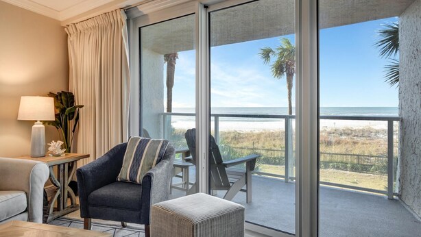 Ein Zimmer mit einer großen SchiebeglasTür, die auf einen Balkon mit Blick auf den Strand und Palmen führt. Es gibt ein Sofa, einen Sessel und einen kleinen Tisch mit Lampe und Dekoration.