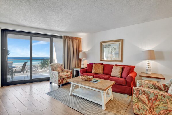 Een woonkamer met een rode bank, een gestreept fauteuil, een salontafel en uitzicht op het strand door grote glazen schuifdeuren.