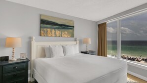 A hotel room with a large bed, two bedside tables with lamps, a painting on the wall, and a view of the beach through a large window. - Gulf Views & Golf Cart: Luxe Seaside Retreat (Miramar Beach)