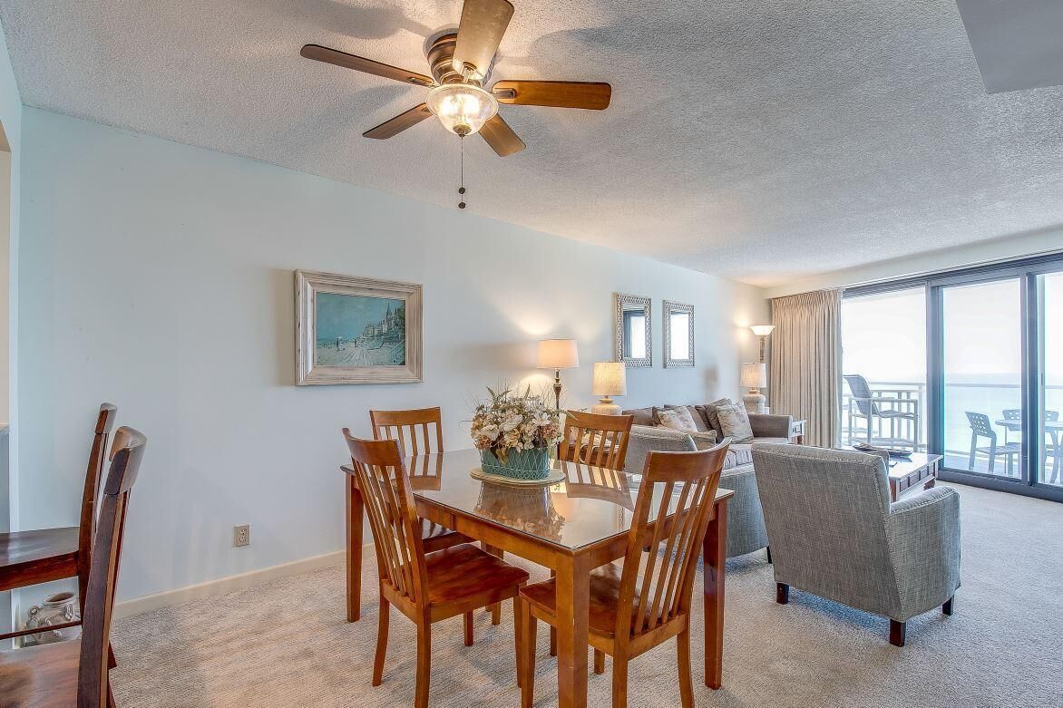 A dining area with a round table, wooden chairs, and a ceiling fan. There is a sofa and a window with a view of the ocean.