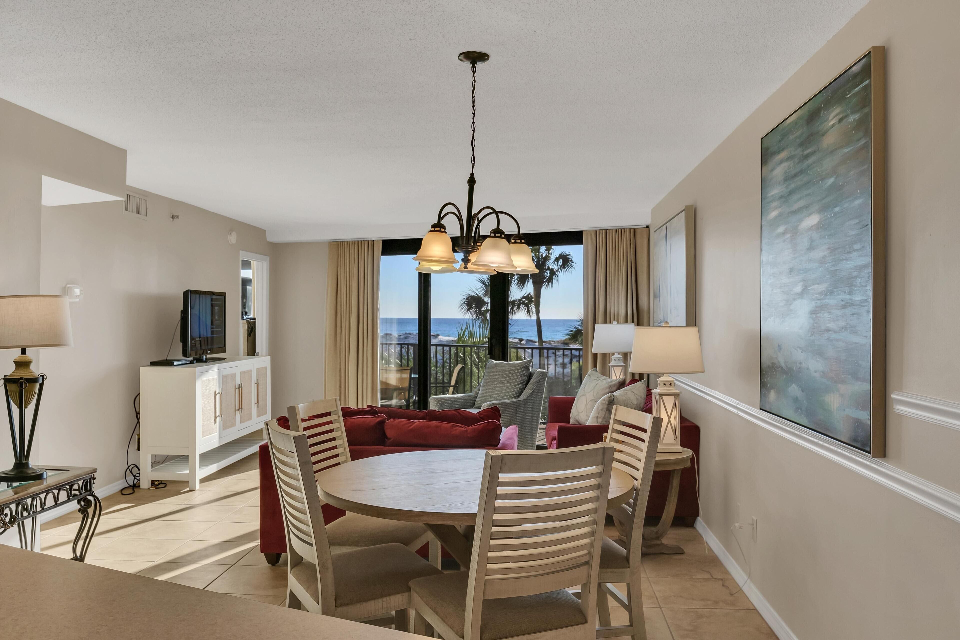 A hotel room with a dining area, a TV on a cabinet, a large window with a view of palm trees and the ocean, and a painting on the wall.