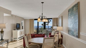 A hotel room with a dining area, a TV on a cabinet, a large window with a view of palm trees and the ocean, and a painting on the wall. - "Sunset Serenity: Recently Renovated 2nd-Floor Oasis with Gulf Views in Beachside One!" at Sandestin Golf and Beach Resort! (Miramar Beach)