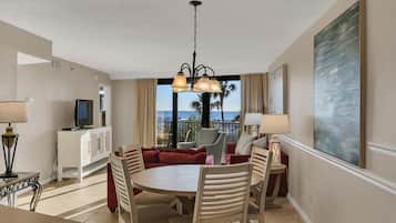 A hotel room with a dining area, a TV on a cabinet, a large window with a view of palm trees and the ocean, and a painting on the wall.