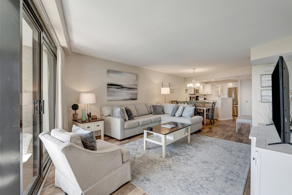 Ein modernes Wohnzimmer mit einem Ecksofa, einem Couchtisch und einem Fernseher. Im Hintergrund ist eine Küche mit Essbereich zu sehen.