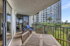 Ein Balkon mit einem Holztisch und Korbstühlen, mit Blick auf einen gepflegten Garten und ein hohes Gebäude.