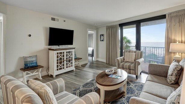 A hotel room with a large TV, a balcony with curtains, and a view of the ocean.