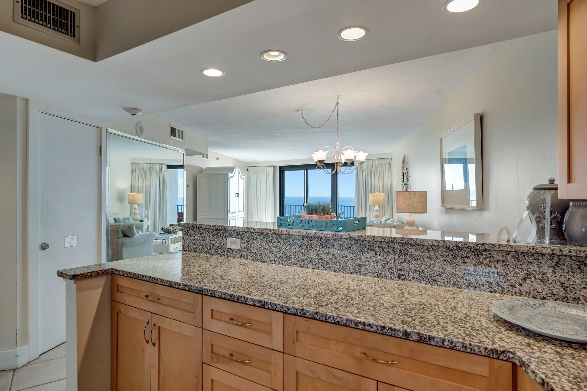 A kitchen with granite countertops, wooden cabinets, and a central island. There is a chandelier above the island and a large window providing a view of the outdoors.