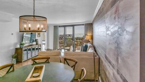 Ein moderner Essbereich mit einem runden Tisch, Stühlen und einer Chandelüster. Es gibt eine Sofagarnitur und einen Blick auf die Stadt durch große Fenster.