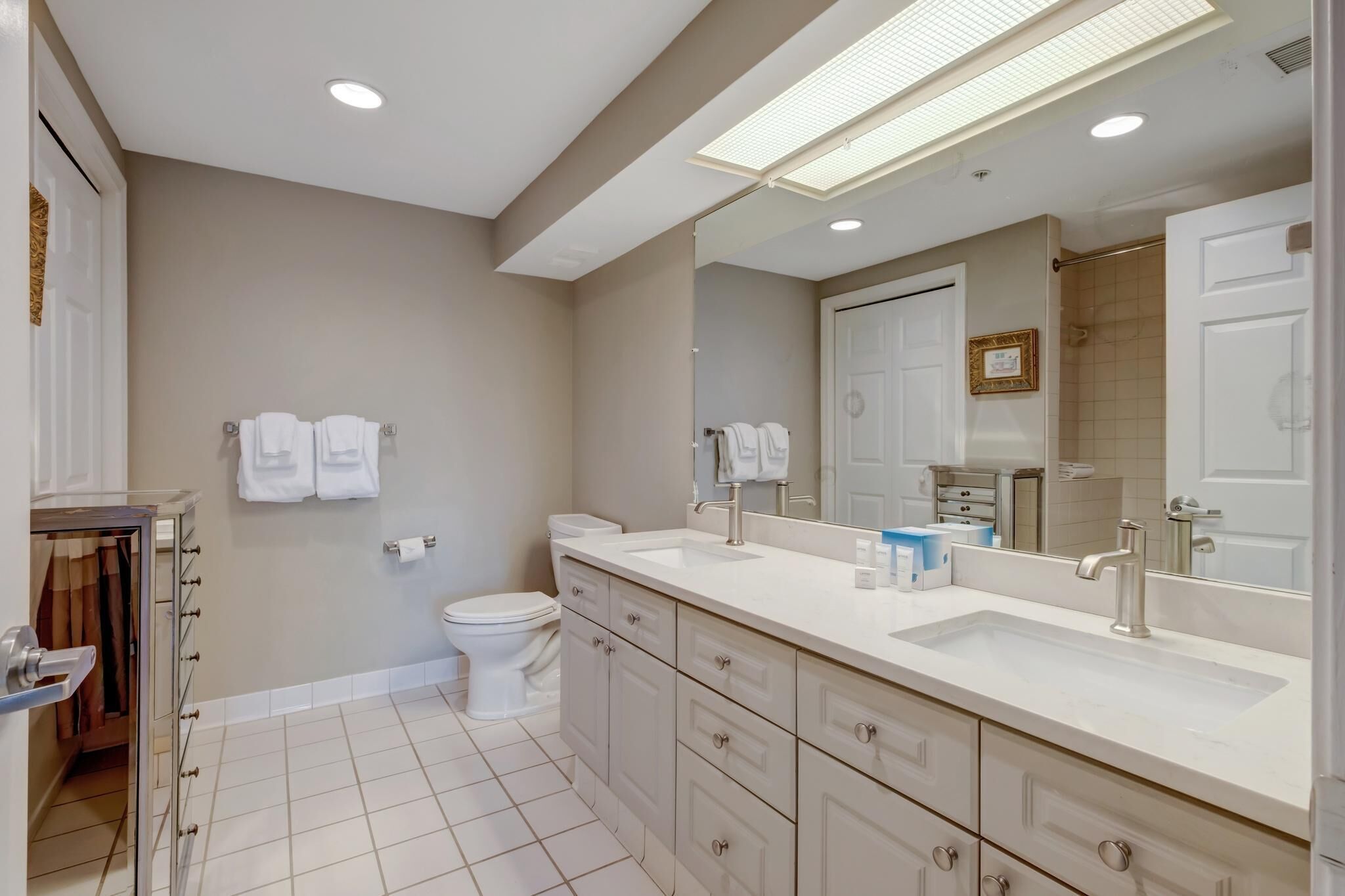 A modern bathroom with a large mirror, two sinks, a toilet, and a walk-in shower.