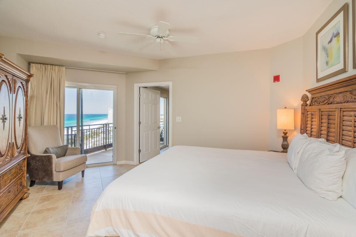 A hotel room with a large bed, a chair, a wooden dresser, a ceiling fan, and a view of the ocean through a sliding door.
