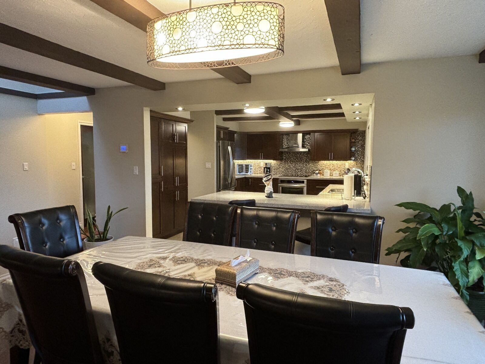 A dining area with a table and chairs, a kitchen in the background, and a hanging light fixture.