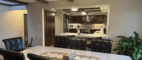 A dining area with a table and chairs, a kitchen in the background, and a hanging light fixture.