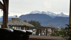 A view from a window overlooking a mountain range with a thatched-roof structure and a gazebo in the foreground. - Mountain View Farm Experience! (Pitt Meadows)