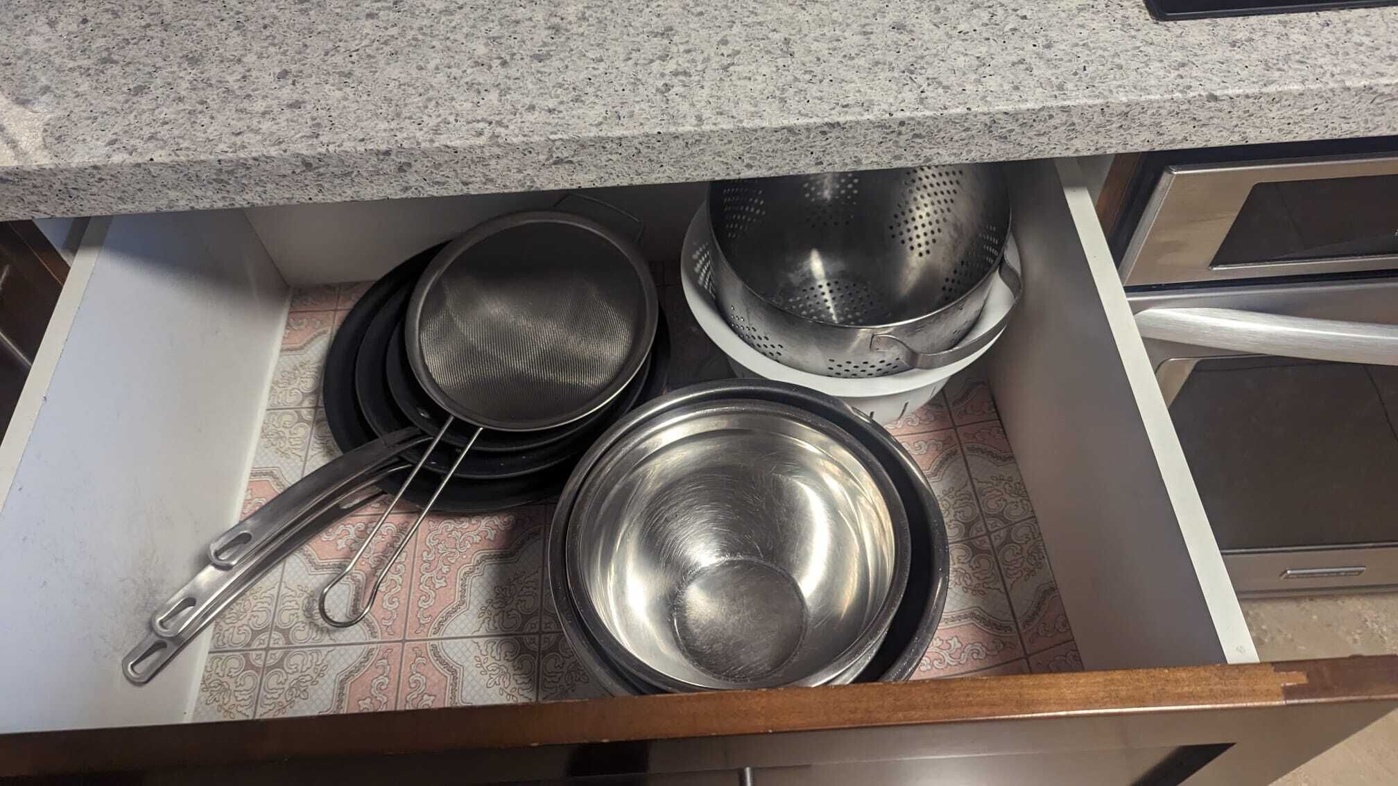 A kitchen drawer containing stainless steel cookware including a pan, a pot, and a colander.