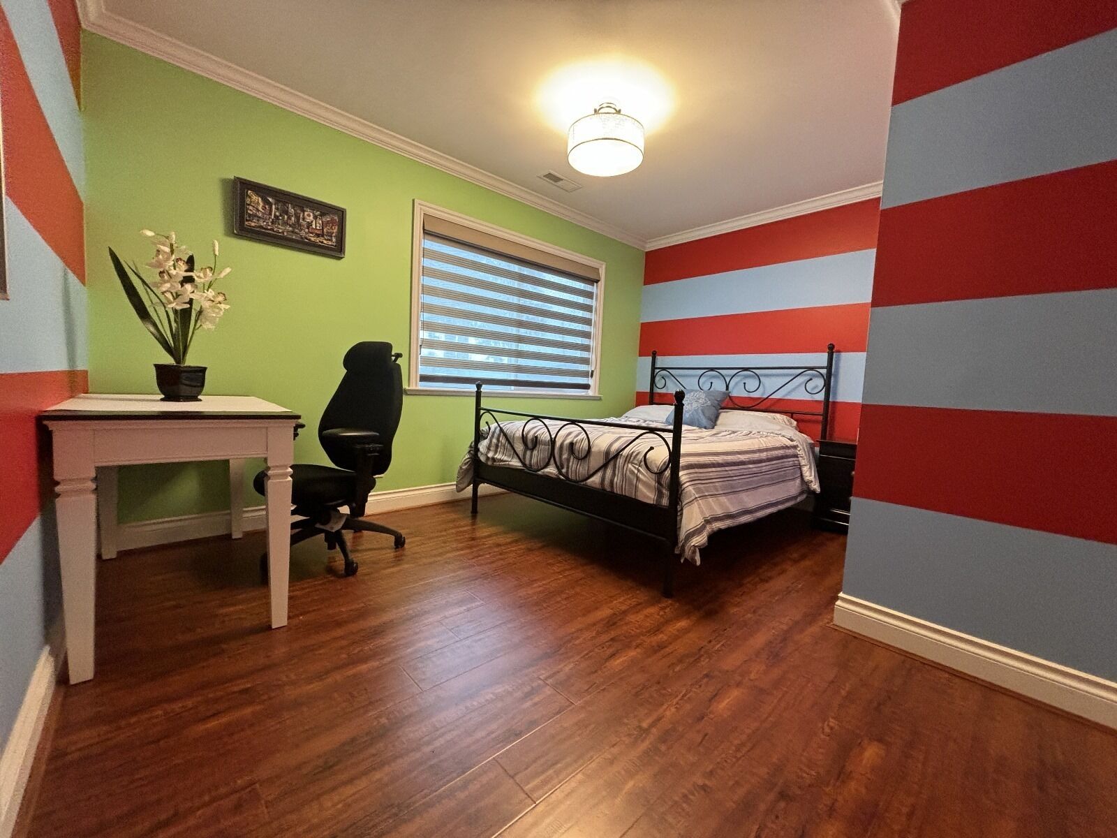 A bedroom with a bed, a desk, a chair, a window with blinds, and a wall with a striped pattern.