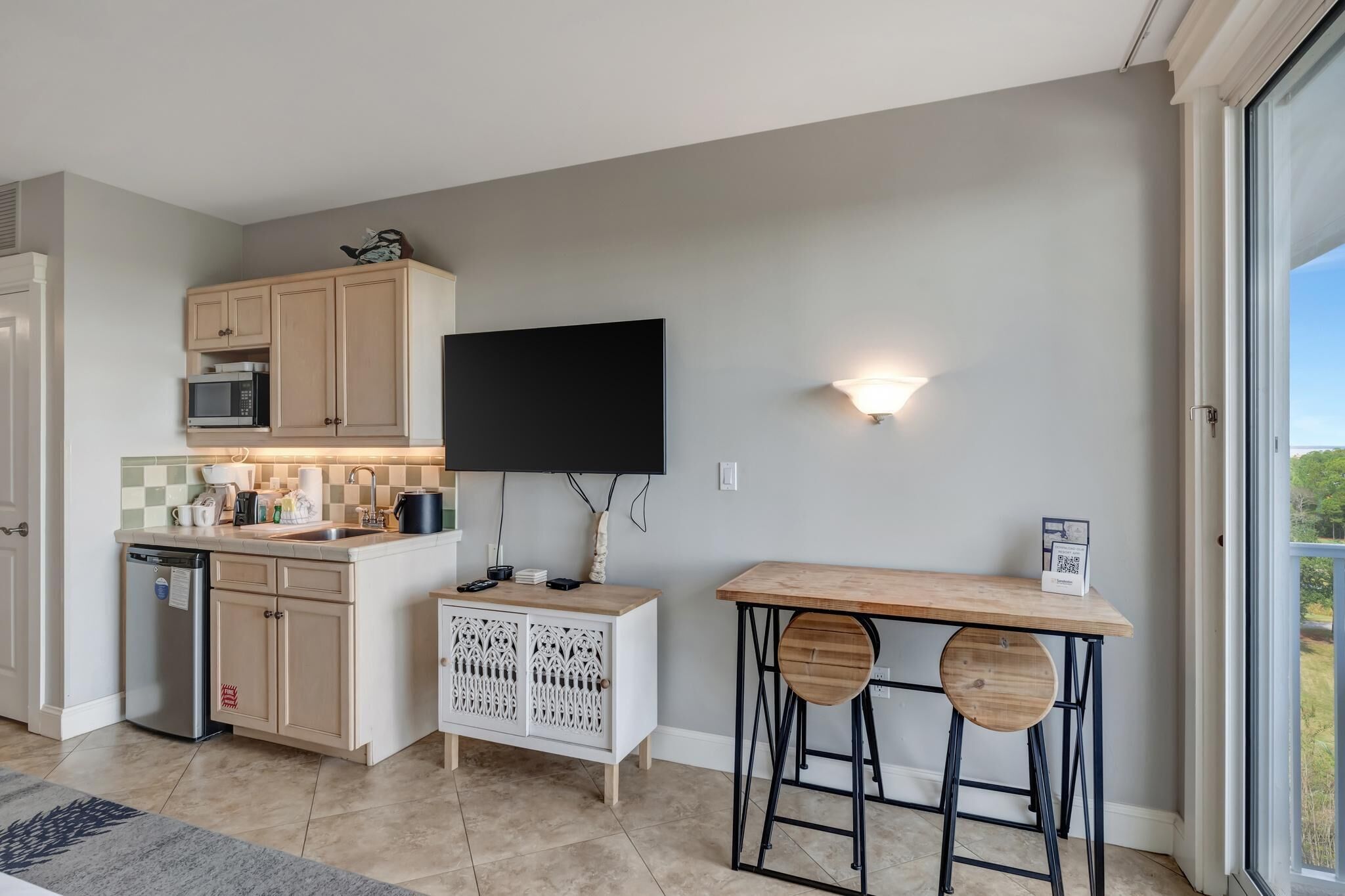 A compact kitchen with a microwave, sink, and cabinets, a flat-screen TV mounted above the counter, and a wooden table with two stools.