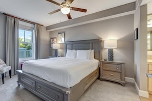 A bedroom with a large bed, a ceiling fan, a nightstand with a lamp, and a window with curtains.
