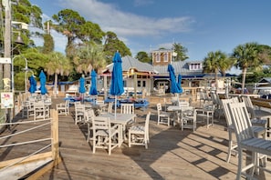 A wooden deck with white tables and chairs, blue umbrellas, and a building in the background. - Serene Choctawhatchee Bay Views from 4th Floor One Bedroom, Two Bathroom Condo with Large Private Balcony at Sandestin Golf and Beach Resort! (Miramar Beach)