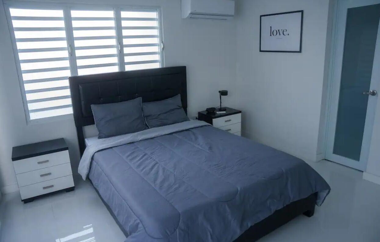 A bedroom with a bed, a nightstand, a drawer, a framed word on the wall, and a door.