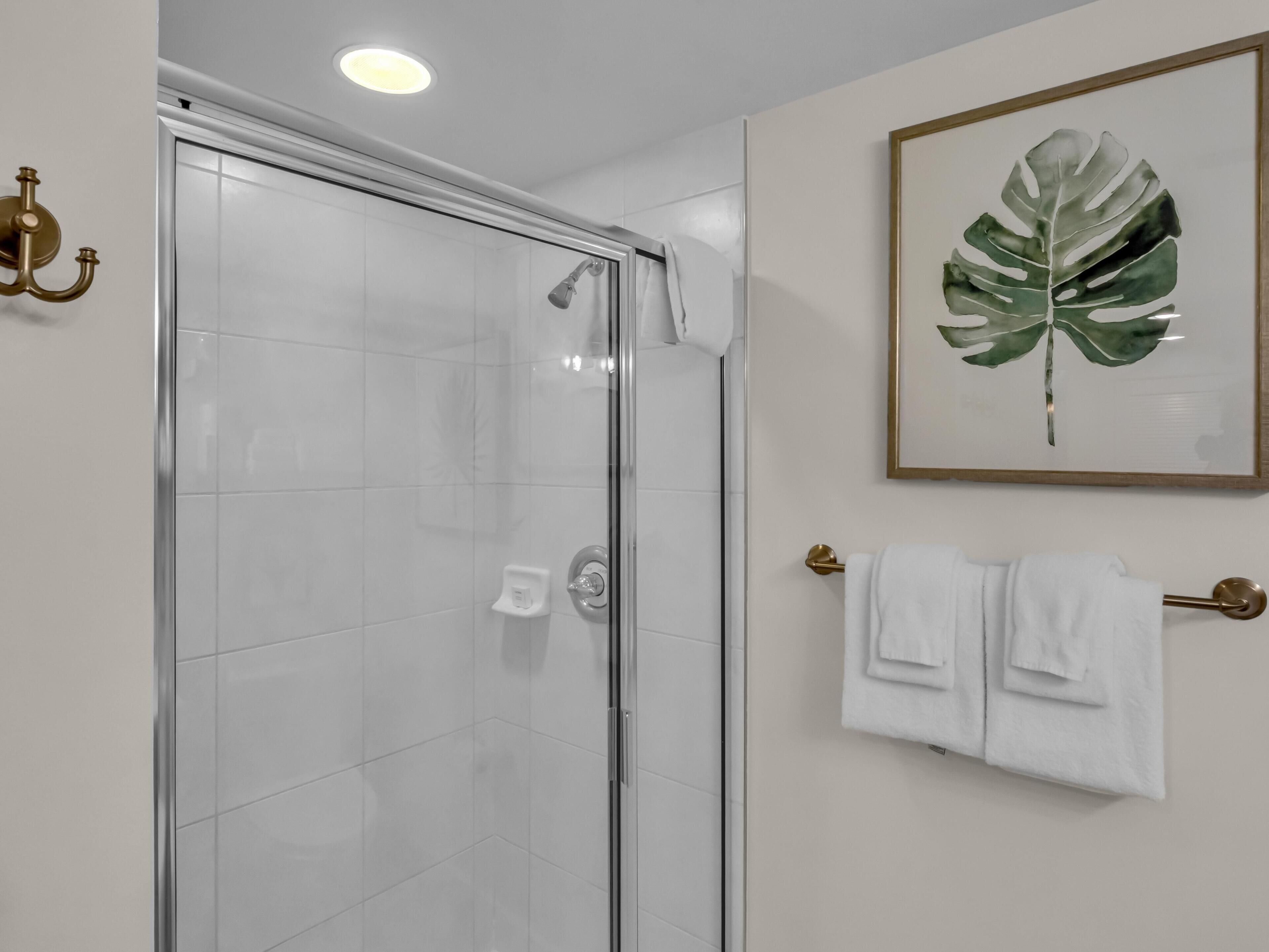 Une salle de bain moderne avec une douche à parois de verre, une œuvre d’art représentant des feuilles encadrée au mur et un porte-serviettes avec des serviettes pliées.