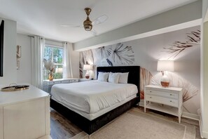 Un dormitorio con cama, mesitas de noche, ventilador de techo y ventana con cortinas.