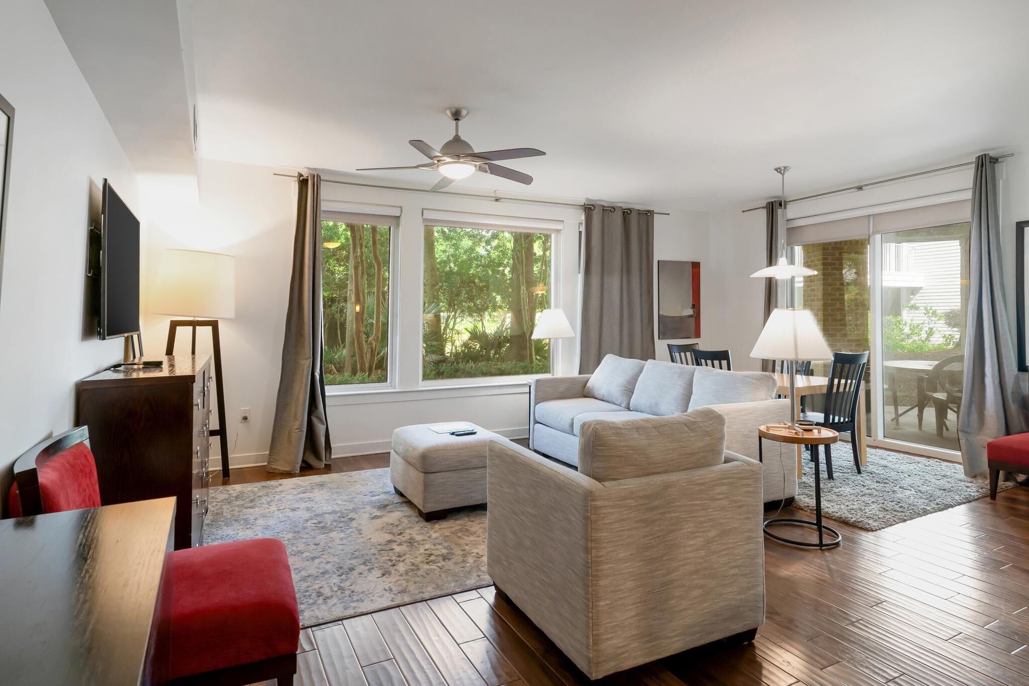 En moderne stue med sofa, skammel og spisebord med stole. Der er en ventilator i loftet, et fjernsyn ophængt på væggen og et vindue med gardiner.