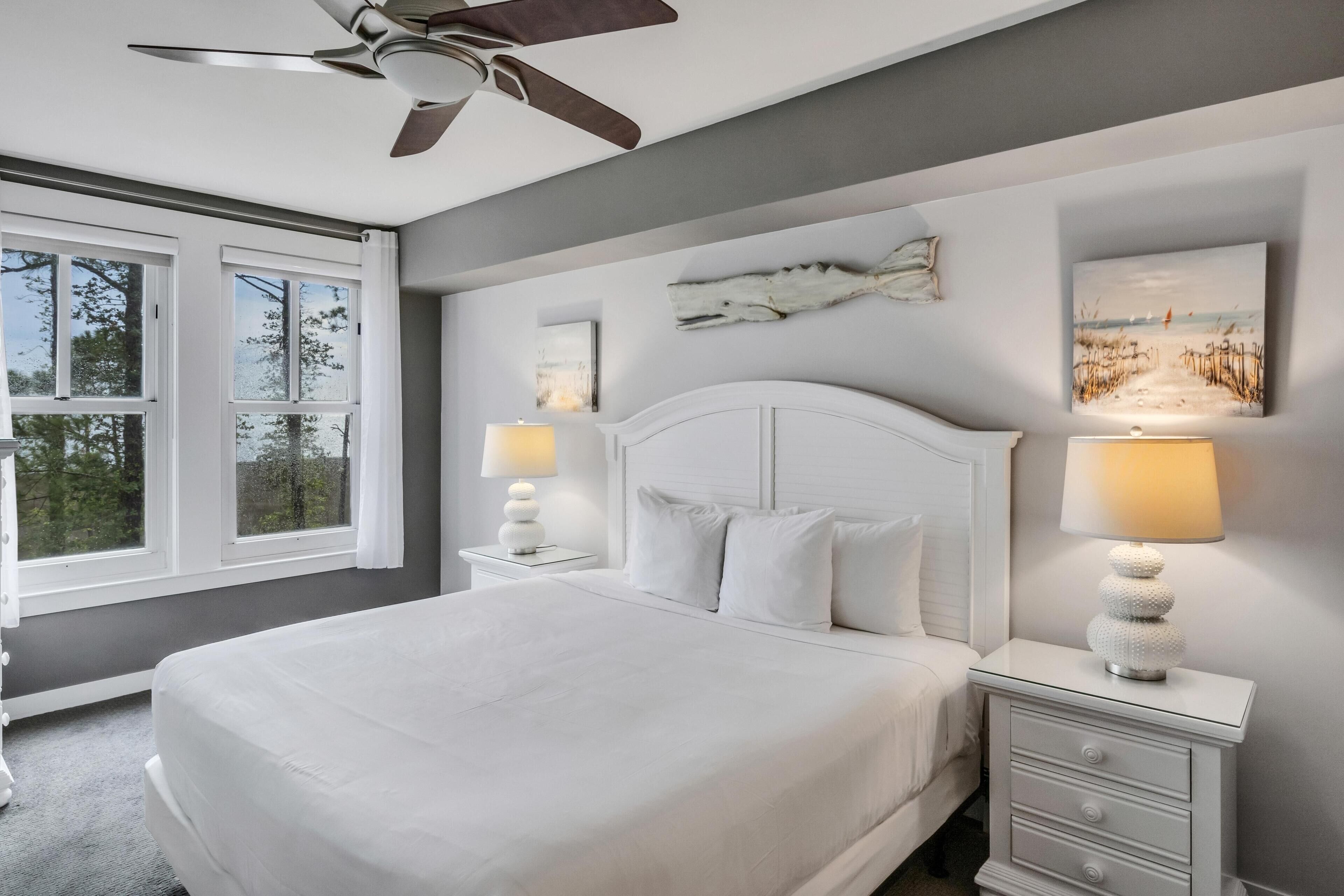 A bedroom with a large bed, two bedside tables with lamps, a ceiling fan, and artwork on the walls.