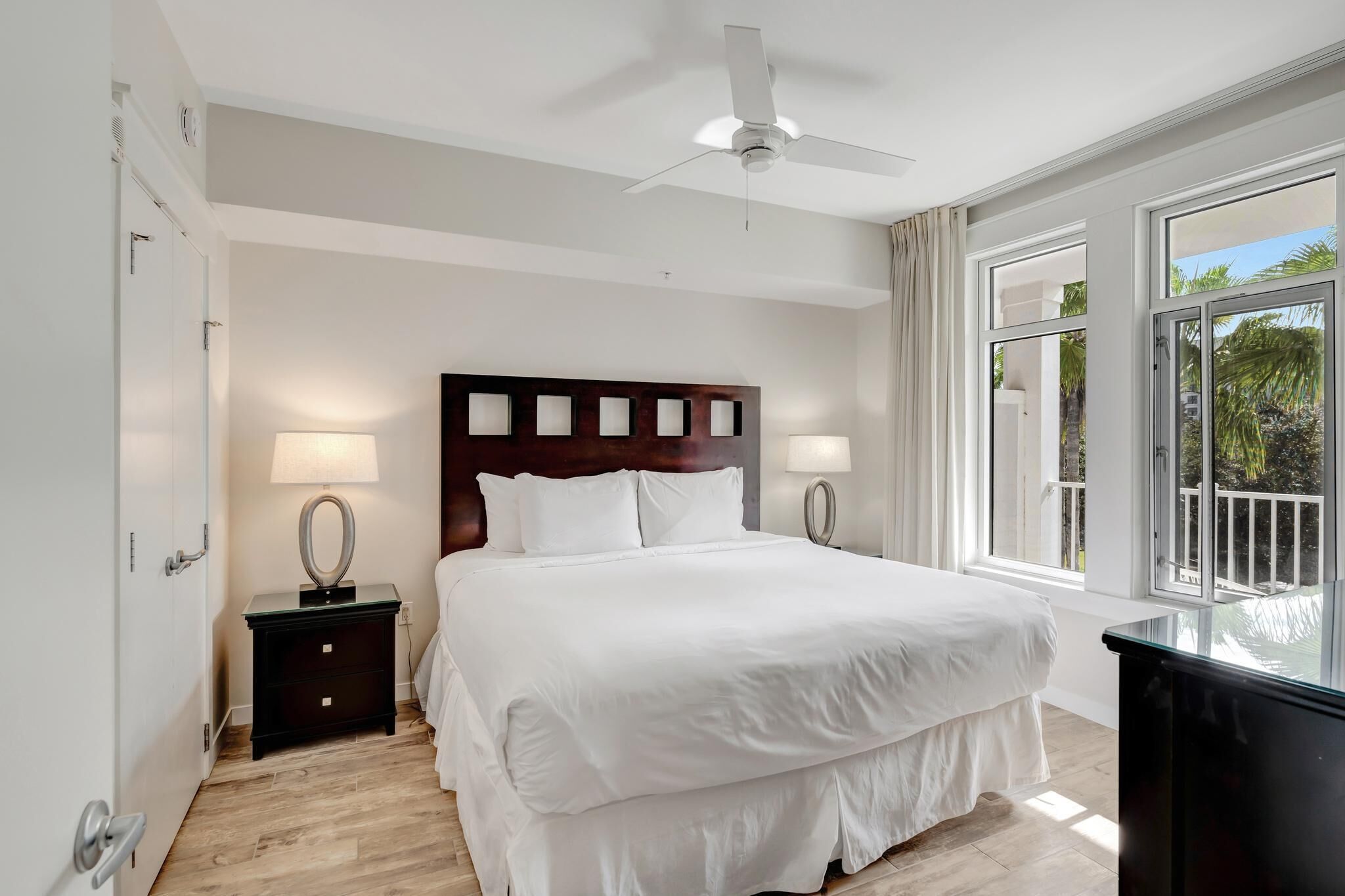 Ein modernes Hotelzimmer mit einem großen Bett, zwei Nachttischen, einem Deckenventilator und einem großen Fenster mit Blick auf Palmen.