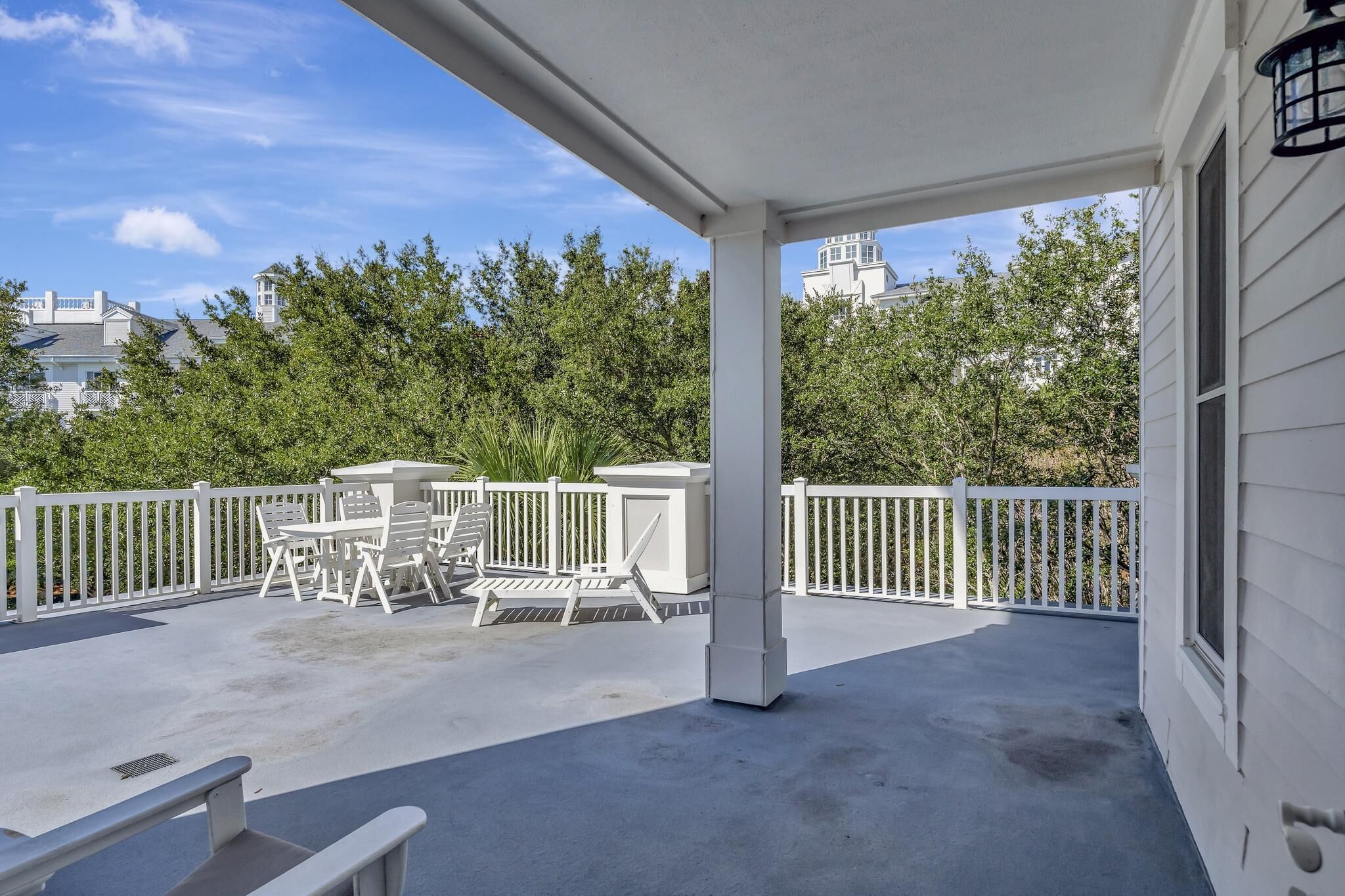A covered patio with white railings, a table and chairs, and a view of trees and buildings in the background.