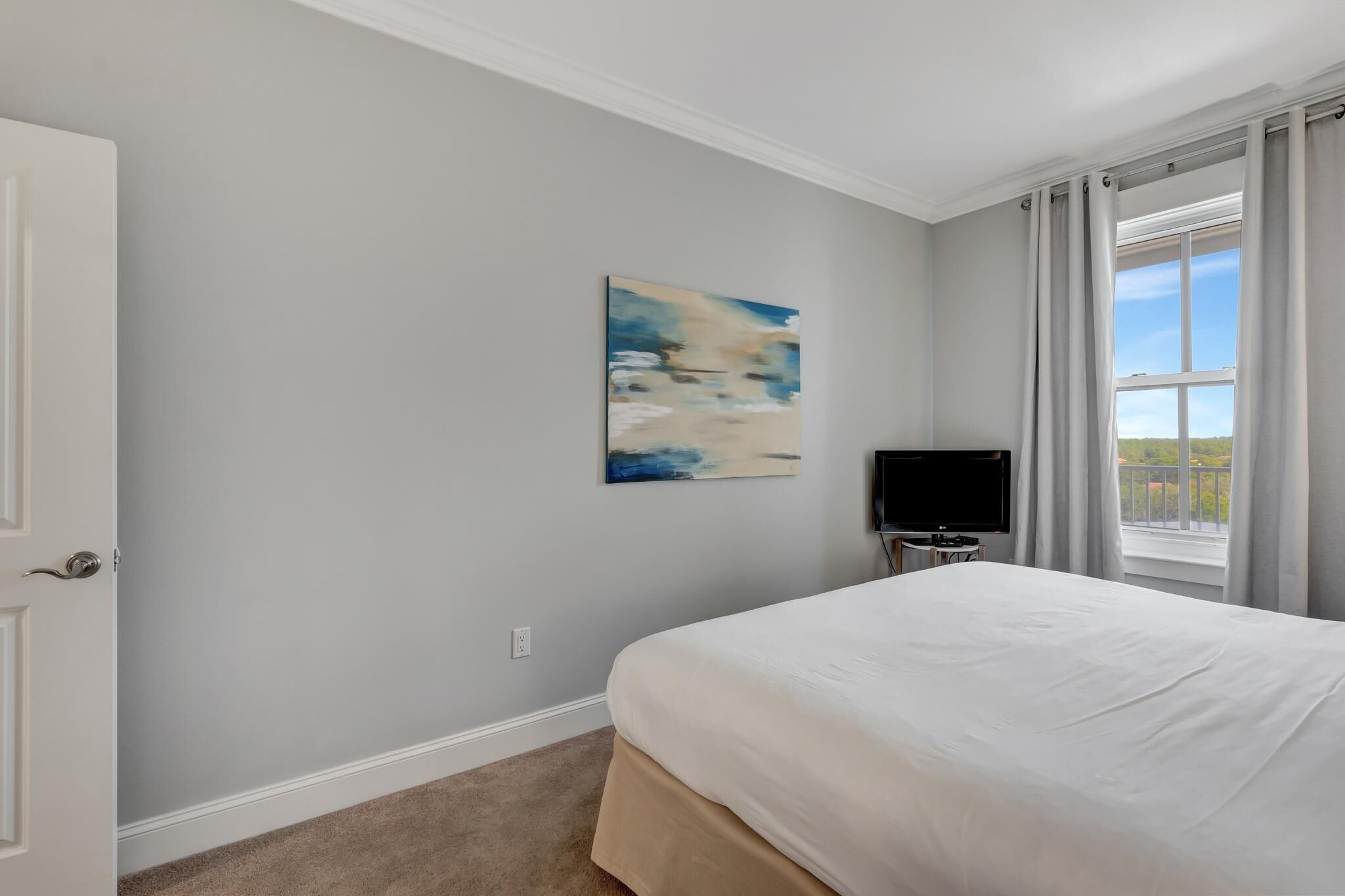 Een slaapkamer met een bed, een schilderij aan de muur, een televisie op een standaard en een raam met gordijnen.