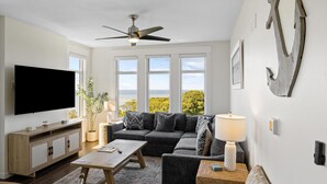 A living room with a large flat-screen TV, a ceiling fan, a sectional sofa, a coffee table, and a view of the ocean through large windows.