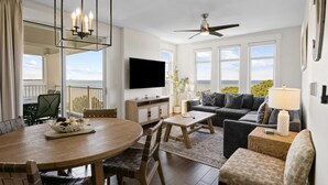 A spacious living room with a large window, a ceiling fan, a flat-screen TV, and a dining area with a wooden table and chairs.