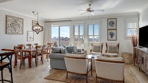 Un salon spacieux comprenant un coin repas, de grandes fenêtres, un ventilateur de plafond et un téléviseur posé sur un meuble.
