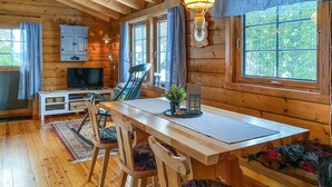 Ein gemütliches Holzhaus-Interieur mit Esstisch, Stühlen, Fernseher und einem Fenster mit Blick auf Bäume.