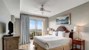 A hotel room with a large bed, two bedside tables with lamps, a ceiling fan, a television on a cabinet, and a view of the outdoors through a sliding door.
