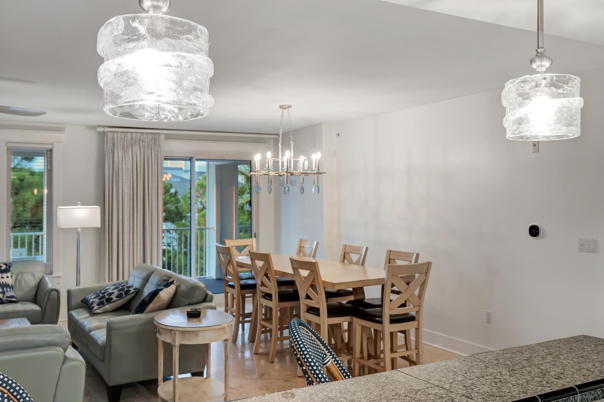 A modern dining area with a wooden table and chairs, a chandelier, and a view of greenery outside.