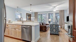 A modern kitchen with a central island, a dining area with a sofa and patterned armchair, and large windows offering a view of trees. - Pilot House One-Bedroom Charm - Your Relaxing Retreat in the Heart of Sandestin at Sandestin Golf and Beach Resort! (Miramar Beach)