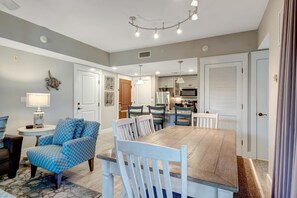 A dining area with a wooden table and chairs, a round side table with a lamp, and a blue patterned armchair. - Pilot House Village One-Bedroom Gem - Your Upscale Getaway at Sandestin Golf and Beach Resort! (Miramar Beach)