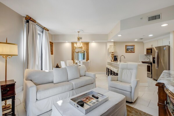 Ein modernes Wohnzimmer mit Sofa, Sessel und Couchtisch. Im Hintergrund ist ein Essbereich mit einer Chandeliebre sichtbar.