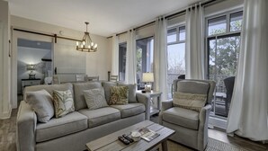 Una sala de estar con sofá, sillón y mesa de centro. Hay una araña, una lámpara y una ventana con cortinas.